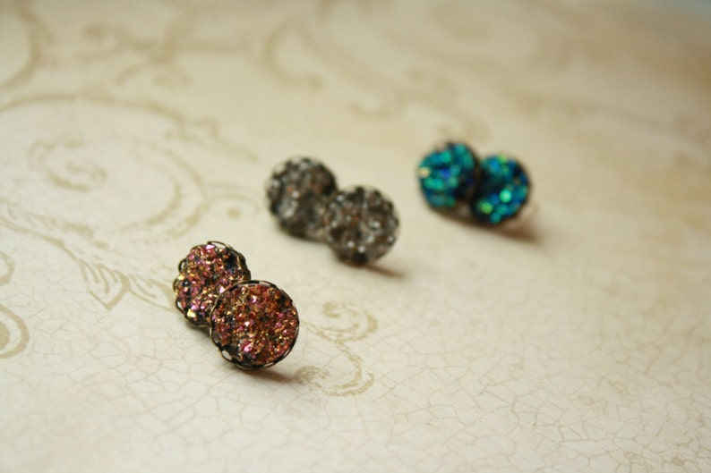 May include: Close-up of three pairs of druzy stud earrings in various colors. One pair is a mix of pink and gold, another is a dark gray, and the last is a vibrant teal. The earrings have a textured, sparkling appearance.