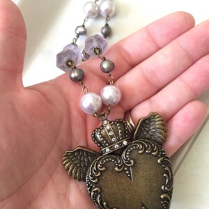 May include: A vintage-style necklace featuring a bronze heart pendant with a crown and wings design. The necklace includes a beaded chain with pearls and faceted beads in shades of grey and white. The pendant is approximately 5 cm in length.