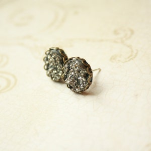May include: A pair of stud earrings with a silver-colored, textured center and a dark bronze-colored, scalloped edge. The earrings are set against a cream-colored background.