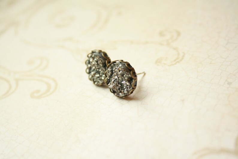May include: A pair of stud earrings with a silver-colored, textured center and a dark bronze-colored, scalloped edge. The earrings are set against a cream-colored background.