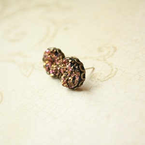 May include: A pair of stud earrings with a dark metal setting. The earrings feature a cluster of iridescent, pink, and gold crystals. The earrings are round and have a post backing. The background is a light, textured surface.