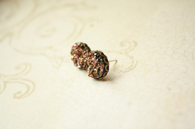 May include: A pair of stud earrings with a dark metal setting. The earrings feature a cluster of iridescent, pink, and gold crystals. The earrings are round and have a post backing. The background is a light, textured surface.