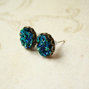 May include: A pair of stud earrings with a round, textured, teal and blue druzy stone set in a bronze-colored metal frame. The earrings have a post backing and are displayed on a cream-colored surface.