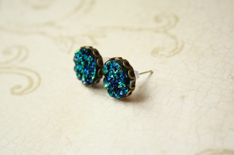 May include: A pair of stud earrings with a round, textured, teal and blue druzy stone set in a bronze-colored metal frame. The earrings have a post backing and are displayed on a cream-colored surface.