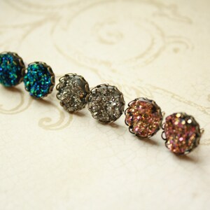 May include: Six druzy stud earrings in a row, each with a different color. The colors include teal, silver, and shades of pink. Each earring has a dark metal filigree setting.