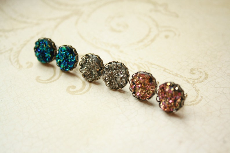 May include: Six druzy stud earrings in a row, each with a different color. The colors include teal, silver, and shades of pink. Each earring has a dark metal filigree setting.