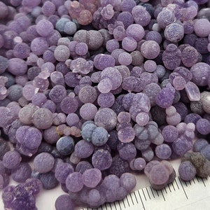 May include: A close-up of a pile of purple agate nodules, also known as 'grape agate', with a ruler for scale. The nodules are various sizes, with some being smaller than a pea and others being larger than a dime. The text 'Medium Size Clusters' is visible in the image.