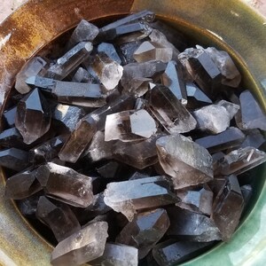 May include: A bowl filled with smoky quartz crystals. The crystals are a dark brown color and have a pointed shape. The bowl is a light brown color with a green glaze.