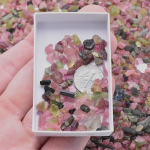 May include: A small white box filled with a variety of pink, green, and black gemstones. The box is sitting on a hand, and a US dime is included for scale.