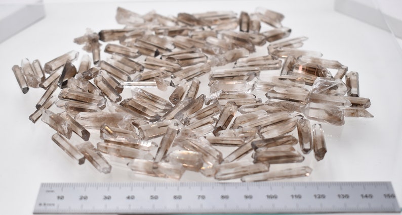 Peut inclure: Un ensemble de pointes de cristal de quartz fum&eacute; brut. Les cristaux, de couleur gris-brun&acirc;tre translucide, varient en taille et en forme. Une r&egrave;gle gradu&eacute;e en centim&egrave;tres est visible au bas de l'image.