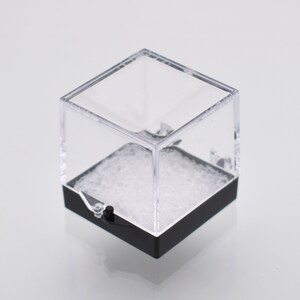 May include: A clear plastic cube display box with a black base. The box is filled with white packing peanuts.