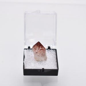 May include: A small, clear quartz crystal with red and white inclusions, displayed in a black and white foam-lined box.