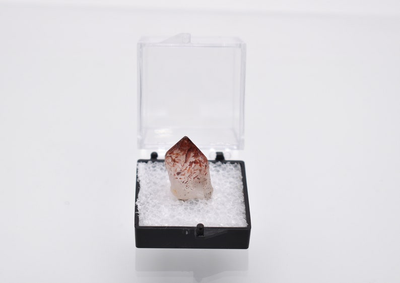 May include: A small, clear quartz crystal with red and white inclusions, displayed in a black and white foam-lined box.