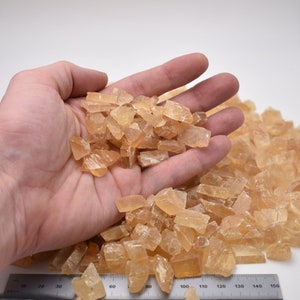 May include: A hand holding a pile of small, translucent, amber-colored crystals. The crystals are scattered on a white surface with a ruler marked in inches.