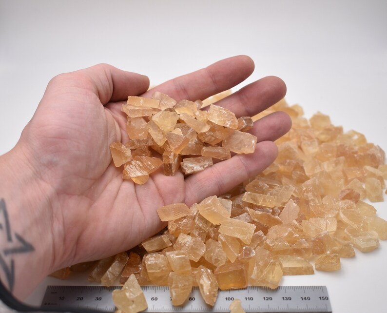 May include: A hand holding a pile of small, translucent, amber-colored crystals. The crystals are scattered on a white surface with a ruler marked in inches.