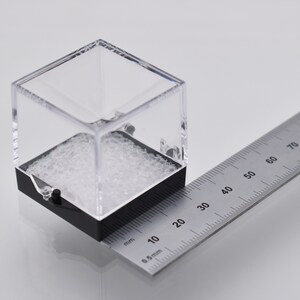 May include: A clear plastic cube with a black base sits on a ruler. The cube measures 2 inches in length. The ruler is marked in millimeters and inches.