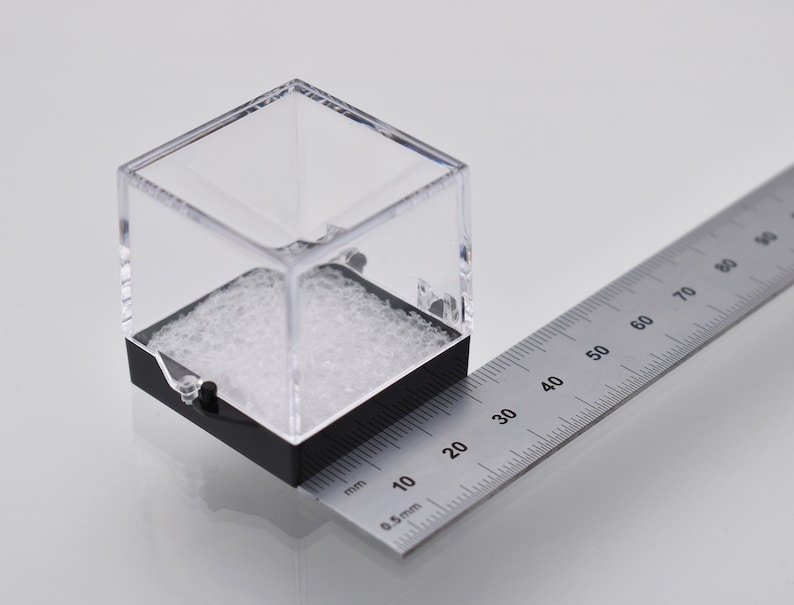 May include: A clear plastic cube with a black base sits on a ruler. The cube measures 2 inches in length. The ruler is marked in millimeters and inches.