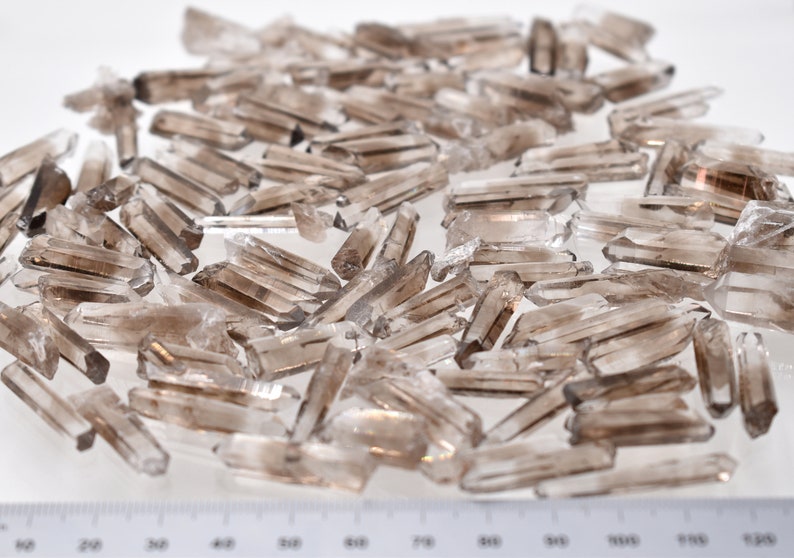 Peut inclure: Une collection de pointes de cristaux de quartz fum&eacute; bruts. Les cristaux varient en taille et en forme, avec une couleur gris-brun&acirc;tre translucide. Ils sont dispers&eacute;s sur une surface blanche, avec une r&egrave;gle visible au premier plan.