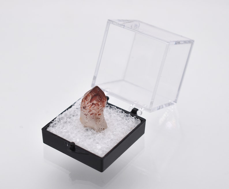 May include: A small, clear quartz crystal with red and brown inclusions is displayed in a black and clear plastic display box.