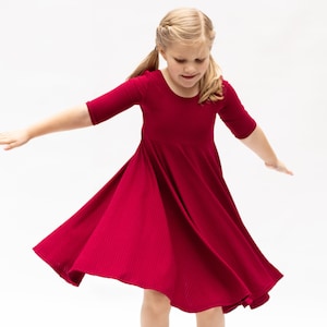 May include: A young girl wearing a red ribbed dress twirls and smiles. The dress has a flared skirt and 3/4 sleeves.