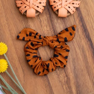 May include: An orange scrunchie with black spots and a bow detail, alongside a pair of orange sandals. The items are arranged on a wooden cutting board, with yellow flowers in the lower left corner.