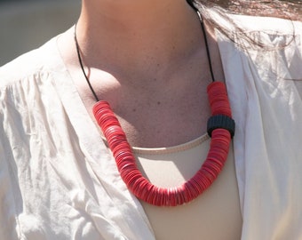 May include: A red disc necklace with a black section. The necklace is worn over a beige top and a cream blouse. The image is taken outdoors in natural light.