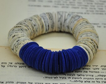 May include: A circular bracelet made from layered paper, with a striking contrast of cream and blue. The central band is a deep blue, and the outer sections display printed text, offering a distinctive, artistic design.