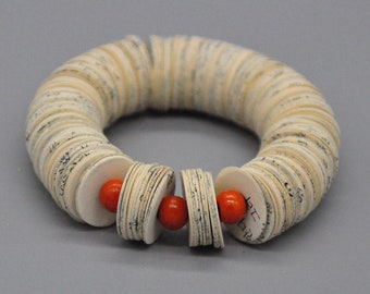 May include: A circular bracelet made of stacked, circular paper pieces with visible text, interspersed with small, round, orange beads. The bracelet has a unique, handcrafted appearance, and is set against a gray background.