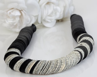 May include: A black and white paper necklace made of layered circles. The circles are made of old book pages and black paper. The necklace is laying on a white surface.