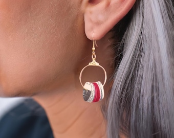 May include: Gold-coloured hoop earrings with a dangling cylindrical charm. The charm features alternating bands of red, silver, and black. The earrings are suspended from a gold-coloured hook.