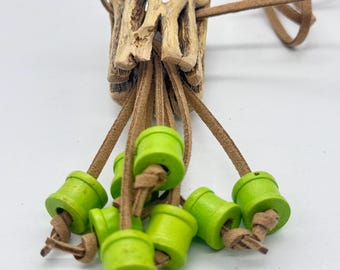Puede incluir: Un collar único con un colgante de madera natural y una serie de cuentas de madera verde. Las cuentas están unidas al colgante con cordones de cuero marrón, creando una estética rústica y terrosa.