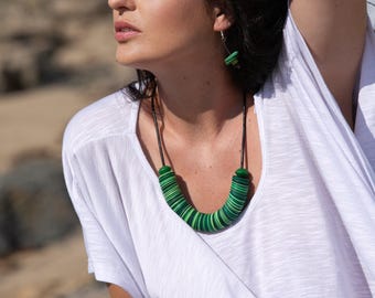 May include: A person wearing a white top and a green and white necklace, earrings, and bracelet. The necklace is made of stacked, circular beads in shades of green. The bracelet is made of similar off-white beads. The earrings are small green squares.