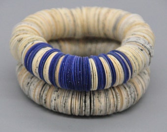 May include: Two circular bracelets, one stacked on top of the other. The bracelets are made of stacked, off-white paper with a section of blue paper. A unique accessory.
