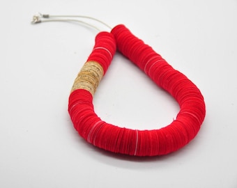 May include: A striking red necklace with a distinctive design. The necklace is composed of numerous small, circular red components, with a section of natural-toned material. It is strung on a thin, silver-coloured wire.