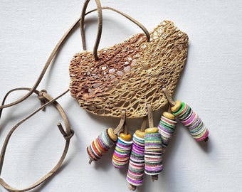 Puede incluir: Un collar con una pieza orgánica grande, porosa y de color tostado suspendida de un cordón de cuero tostado. Cinco dijes cuelgan de la parte inferior, cada uno compuesto por discos delgados, redondos y multicolores en tonos rosa, verde, azul y blanco.