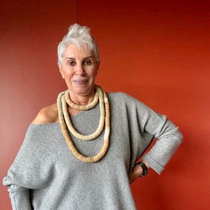 Può includere: Una lunga collana multistrato composta da piccole perline circolari a forma di disco, nei toni del beige e del marrone chiaro. La collana è indossata sopra un maglione grigio chiaro a maniche lunghe, con scollo a barchetta e tessuto morbido.