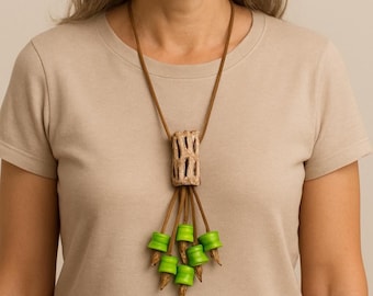 May include: A handmade necklace with a central, textured, beige pendant with vertical openings. Below, brown cords descend, each with a bright green, cylindrical bead and a pointed, brown tip. The necklace is worn against a neutral background.