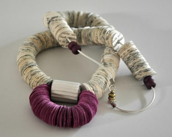 May include: A distinctive necklace with large beads wrapped in paper, in cream and grey tones. It features a bright magenta circular element and a silver clasp. The design incorporates small, dark purple beads and a silver wire.