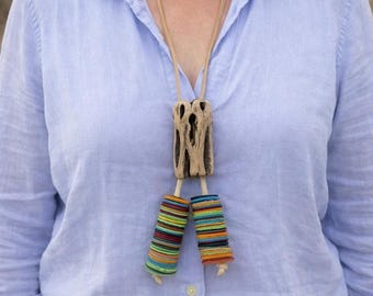 May include: A distinctive necklace with a light brown, rectangular wooden pendant with a textured surface. Below, two cylindrical pendants are wrapped with colourful threads. The necklace hangs from a light brown cord.