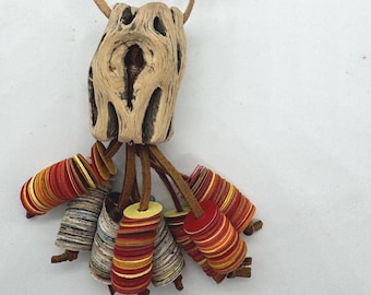 May include: A distinctive necklace with a light brown, carved wooden pendant with multiple holes. Below the pendant hang several cylindrical beads in shades of red, yellow, and grey, strung on brown leather cords. The necklace has a tan leather cord.