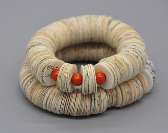 May include: Two stacked bracelets crafted from circular paper pieces, featuring visible text, and interspersed with orange beads. The bracelets are beige and white, with the orange beads providing a vibrant contrast. They rest on a grey background.