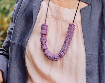 May include: A purple necklace with a U-shaped design. The necklace is made of stacked, circular pieces and connected by a black cord. The necklace is worn over a pink blouse and a blue jacket.