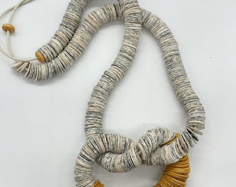 May include: A handmade necklace with a knotted design. The necklace is made of small, round, grey and white beads. A section of the necklace is a golden-yellow colour, creating a striking contrast. The necklace has a cream-coloured cord.