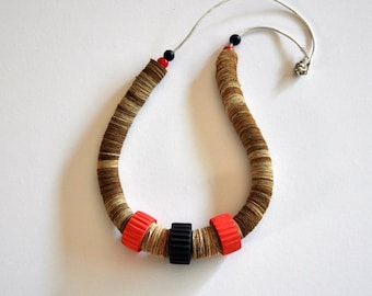 May include: A handcrafted necklace with a brown and beige paper coil design. The necklace features red and black accents, including a black cylindrical bead and two red cylindrical beads. It is strung on a silver-coloured cord.