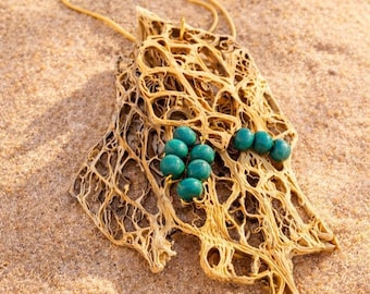 May include: A gold-coloured pendant necklace with a distinctive, organic design. The pendant has a network of interconnected lines and is decorated with several teal-coloured beads. The necklace is displayed on a sandy surface.