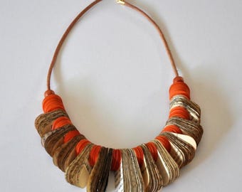 Peut inclure: Un collier composé d'un cordon en cuir marron et d'une série de perles circulaires orange alternant avec des segments de papier plié imprimés de texte noir. Les pièces en papier forment un motif incurvé en éventail sur la partie inférieure.