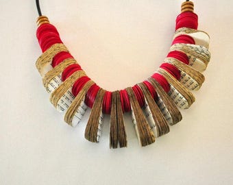 May include: A distinctive necklace crafted from red fabric and folded paper, possibly from a book, creating a layered effect. The necklace is suspended from a black cord. The design is a unique piece of jewellery.