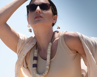 May include: A woman wearing sunglasses, a beige bodysuit, and a light beige shirt. She is also wearing a long necklace with silver and purple beads, and matching earrings. The background is a clear blue sky.
