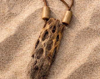 Puede incluir: Un collar con colgante que presenta una pieza de madera de cholla texturizada y porosa suspendida de un cordón de cuero marrón. El collar incluye dos cuentas de madera cilíndricas que aseguran el cordón sobre la estructura de madera orgánica.