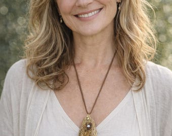 May include: A woman wearing a beige V-neck shirt and a light cardigan. A brown cord necklace with a unique pendant featuring a woven design and a small gemstone is the focal point. The background is blurred with green tones.